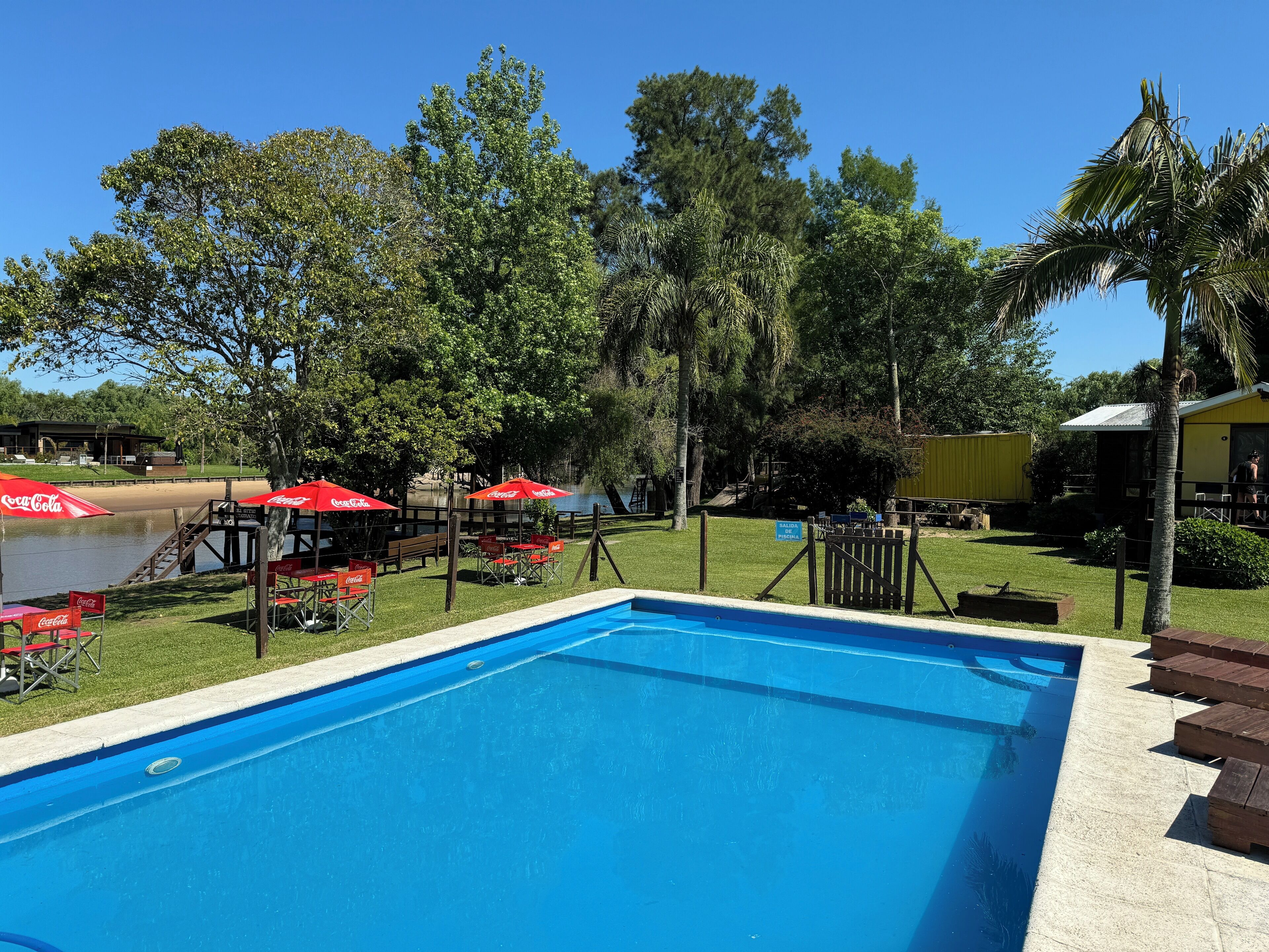 Una piscina con sombrillas rojas con la marca Coca-Cola, rodeada de árboles y un pequeño edificio.