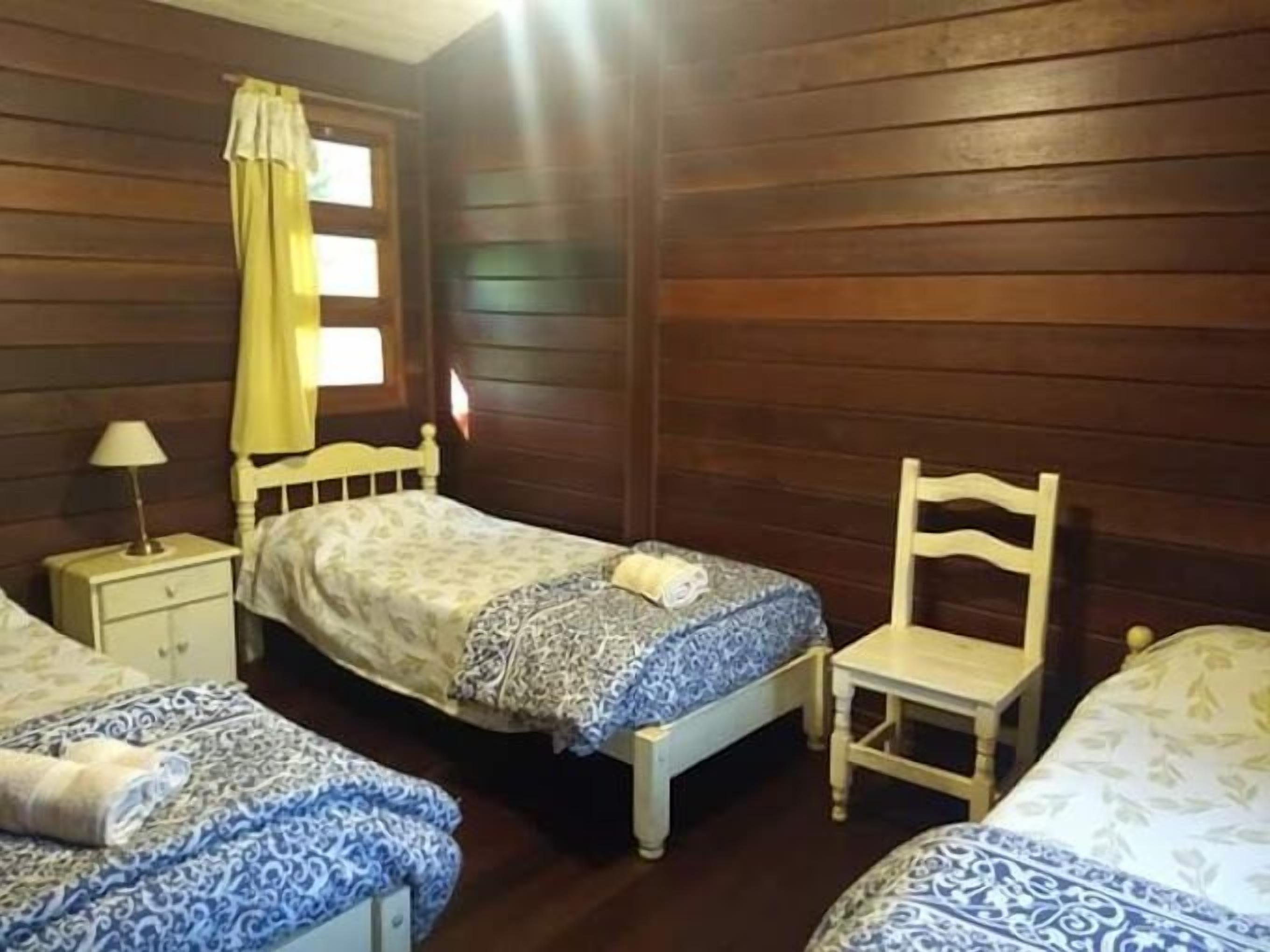 Una habitación con paredes de madera, dos camas con ropa de cama estampada, una mesita de noche con lámpara y una silla.
