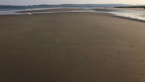 Une vaste plage de sable fin, avec des vagues douces, un ciel dégagé et des collines à l’horizon.