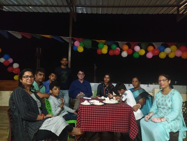 A group of people seated around a table with a red and black checkered tablecloth, under a roofed area with colorful balloons.