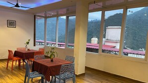 A dining area with a table set for four, surrounded by chairs, large windows with a view of a mountainous landscape, and a ceiling fan. - Hotel The Shivaay - Delight Inn  (Rishikesh)