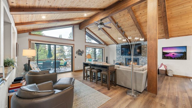 A spacious living room with a stone fireplace, wooden ceiling beams, and a large window overlooking a deck. - Edgewater Escape in Dollar Point Tahoe City (Tahoe City)