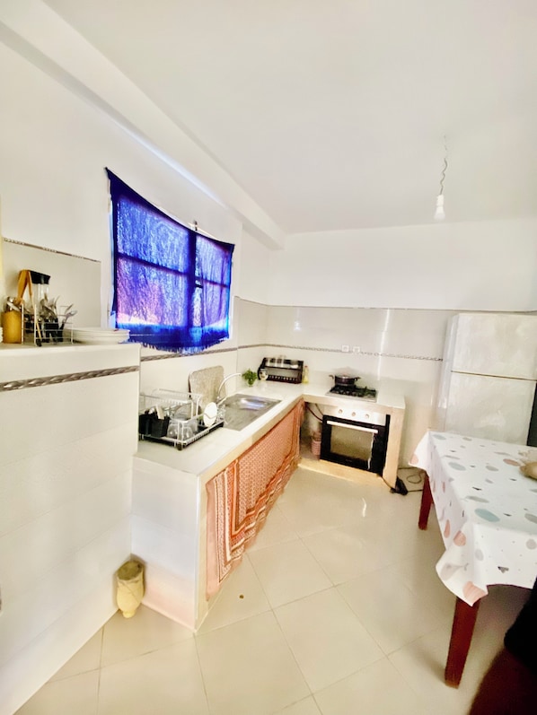 A compact kitchen with a central island, a small dining table, and a window with blue curtains.
