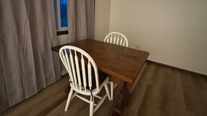 Un coin repas avec une table en bois et deux chaises blanches, une fenêtre avec des rideaux et un mur sobre.