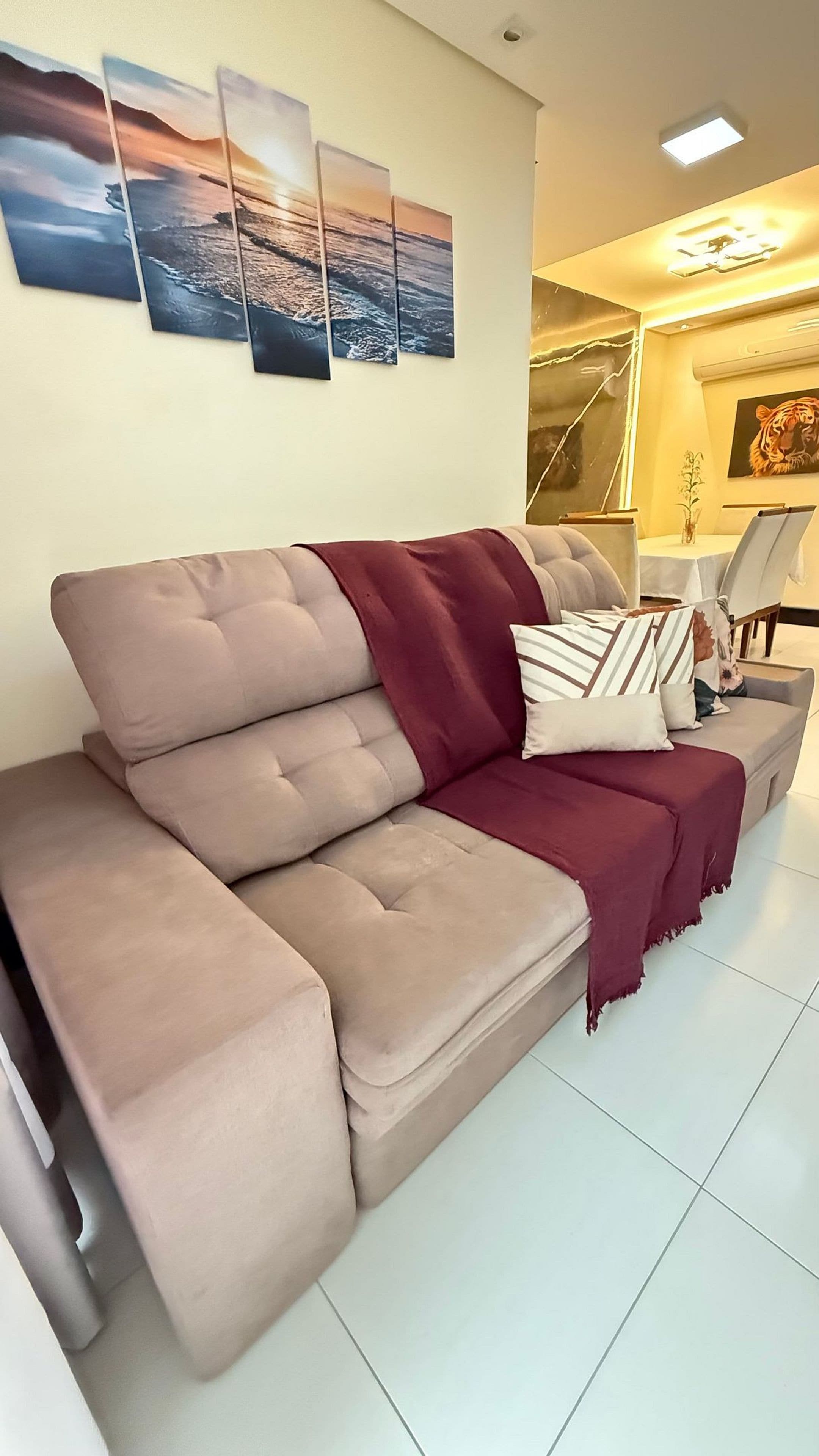 A beige sofa with a maroon throw blanket and pillows, a white table with a vase, and a tiger painting on the wall.