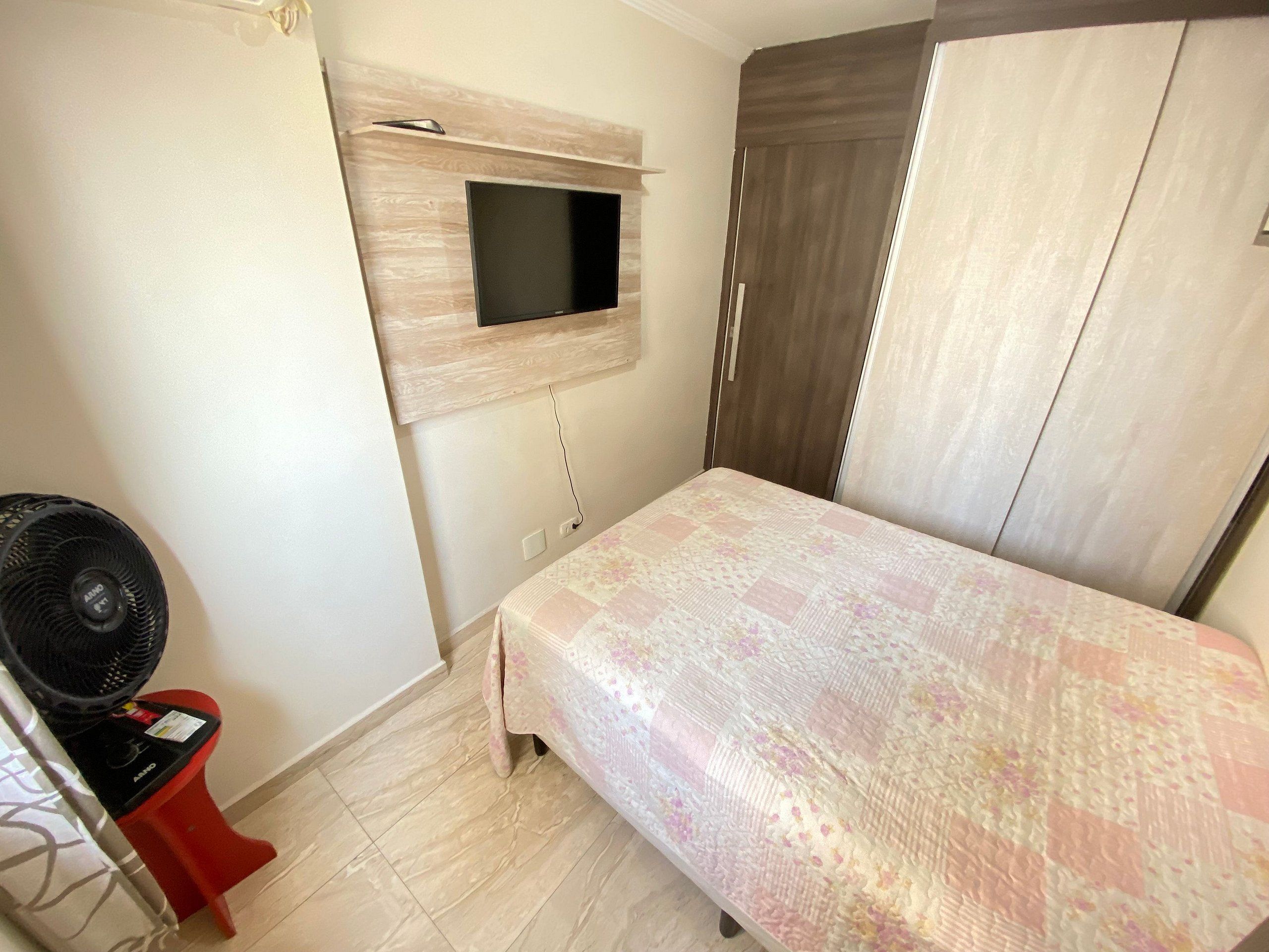 Une chambre à coucher avec un lit, un téléviseur fixé au mur, un fauteuil rouge et un ventilateur.