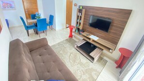 Un saló amb un sofà marró, una televisió penjada a una paret de fusta, una zona de dinar amb cadires blaves i una taula vermella.