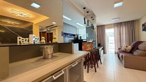 A modern kitchen with a bar area, a dining space with stools, and a living area with a sofa and a TV.