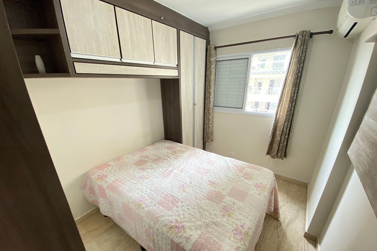 Una habitación pequeña con cama individual, cabecera de madera, ventana con cortinas y suelo de baldosas.