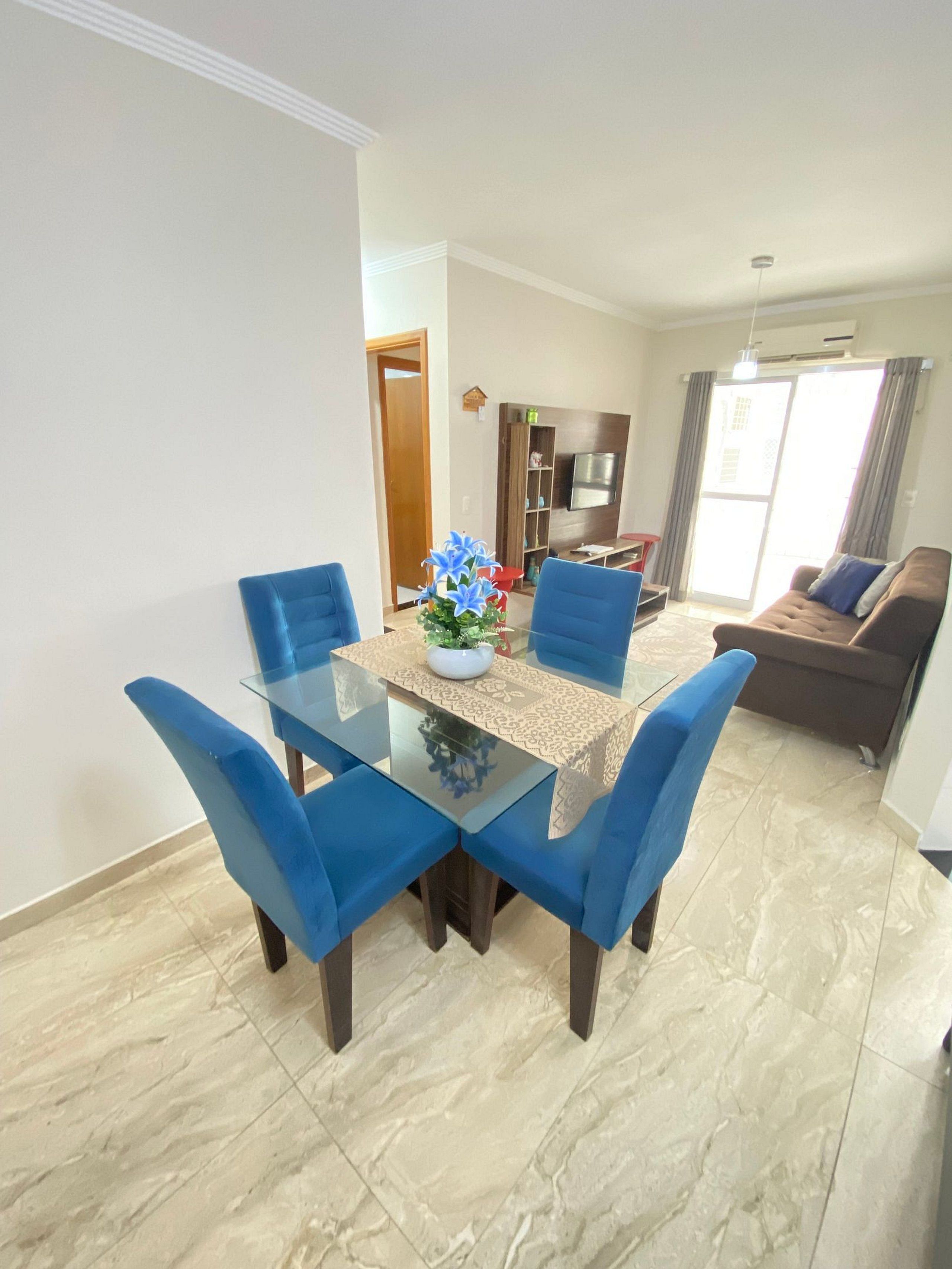 A dining area with a glass table and blue chairs, a sofa in the background, and a large window with curtains.