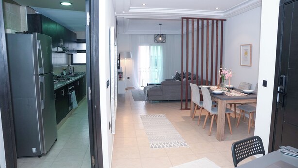 A modern kitchen with a stainless steel refrigerator, a dining area with a wooden table and chairs, and a living room with a sofa and a decorative partition. - Modern and chic apartment | Rabat city center (Rabat)