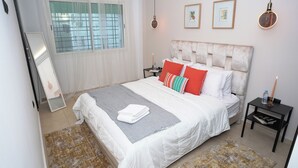 A neatly made bed with white bedding and colorful pillows, a bedside table with a water bottle, and a wall-mounted light fixture. - Modern and chic apartment | Rabat city center (Rabat)