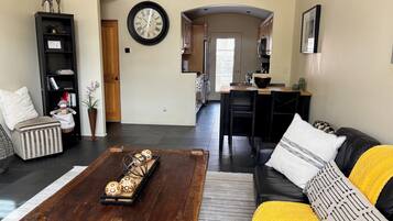 Una sala de estar con una mesa de centro, sofá y estantería. Se ve una cocina al fondo.