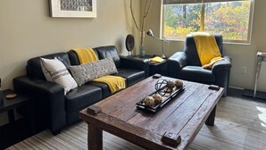 Un salon avec un canapé en cuir noir, une table basse en bois et une fenêtre équipée de stores.