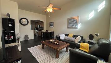 A living room with a dark floor, a wooden coffee table, a black leather sofa with yellow throw pillows, a black bookshelf, and a ceiling fan.