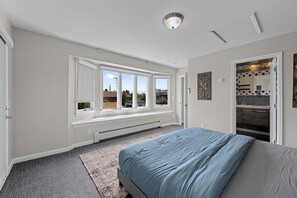 A bedroom with a large bed, a window seat, a view of the outside, and a kitchen area visible through an open door. - Group Retreat with Hot Tub, Gym, Sauna, and Fire Pit (Englewood)