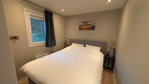 Un dormitorio con una cama grande, una ventana con cortinas, un radiador, una mesita de noche con un florero y un cuadro en la pared.