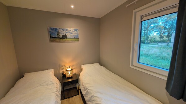 Un dormitorio con dos camas, una mesita de noche con lámpara, un cuadro en la pared y una ventana con vistas a árboles.