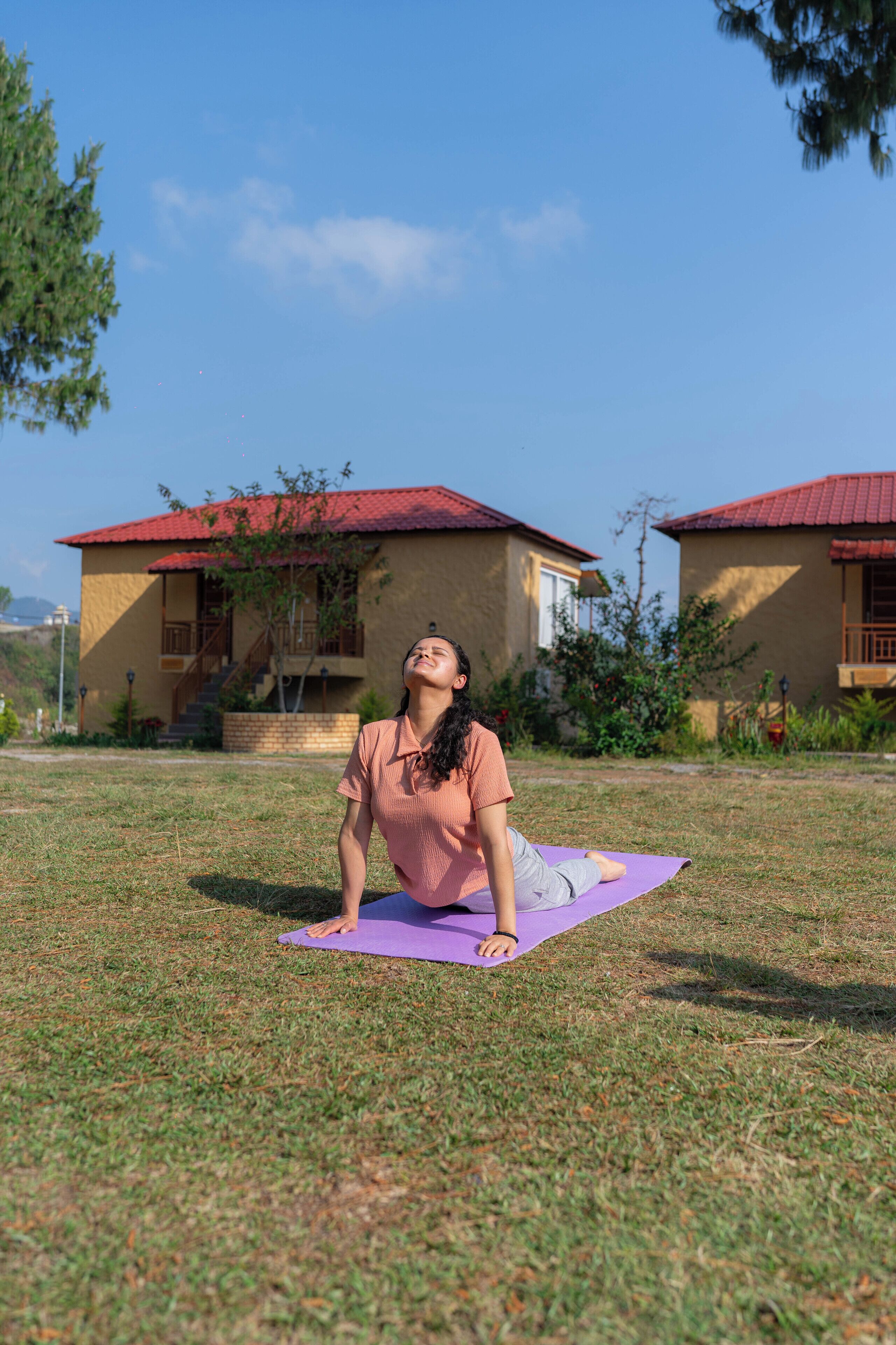 A person performing yoga on a purple mat in a grassy area with buildings in the background.