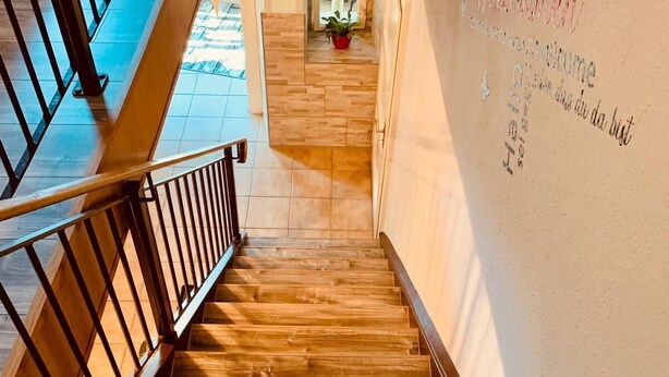 A wooden staircase with a metal railing leading to a landing. A wall with text and a potted plant is visible in the background.