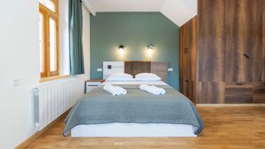 A neatly made bed with a grey quilt, two bedside tables with lamps, a wooden wardrobe, and a window with curtains.