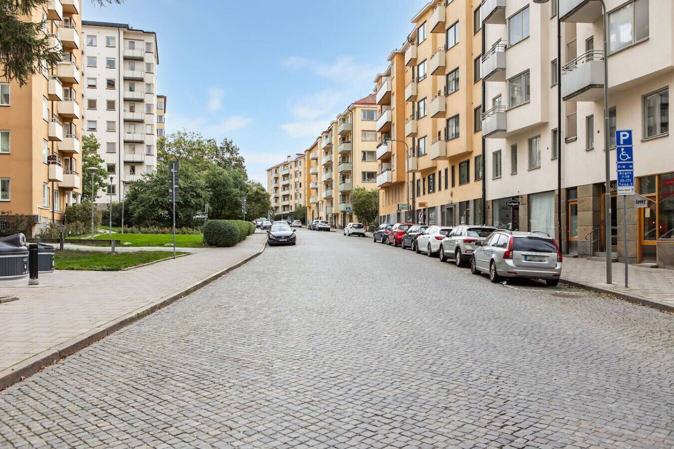 En bostadsgata med parkerade bilar, hyreshus och en klar himmel.
