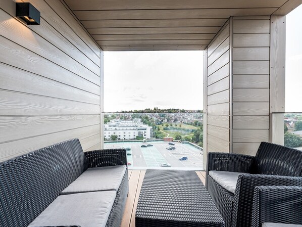 A balcony with wicker furniture, a view of a parking lot, and a building in the background.