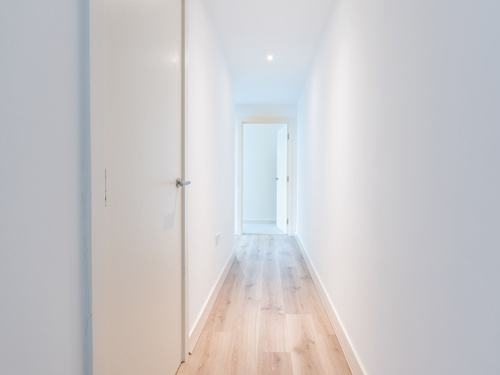 Passadís estret i ben il·luminat amb parets blanques i sòl de fusta, que condueix a una porta al final.
