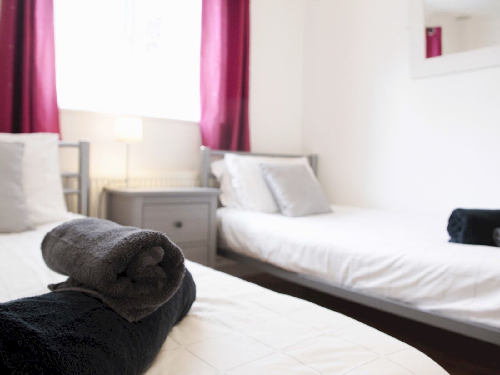 Quarto de hotel com duas camas, roupa de cama branca, mesa de cabeceira com abajur e janela com cortinas.
