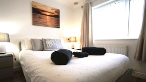 Una cama bien hecha con edredón blanco y dos almohadas, una mesita de noche con lámpara y un cuadro de una puesta de sol en la pared.