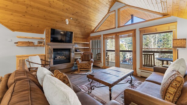 Interior acogedor de una cabaña con techo de madera, chimenea, sofá y ventanas amplias.