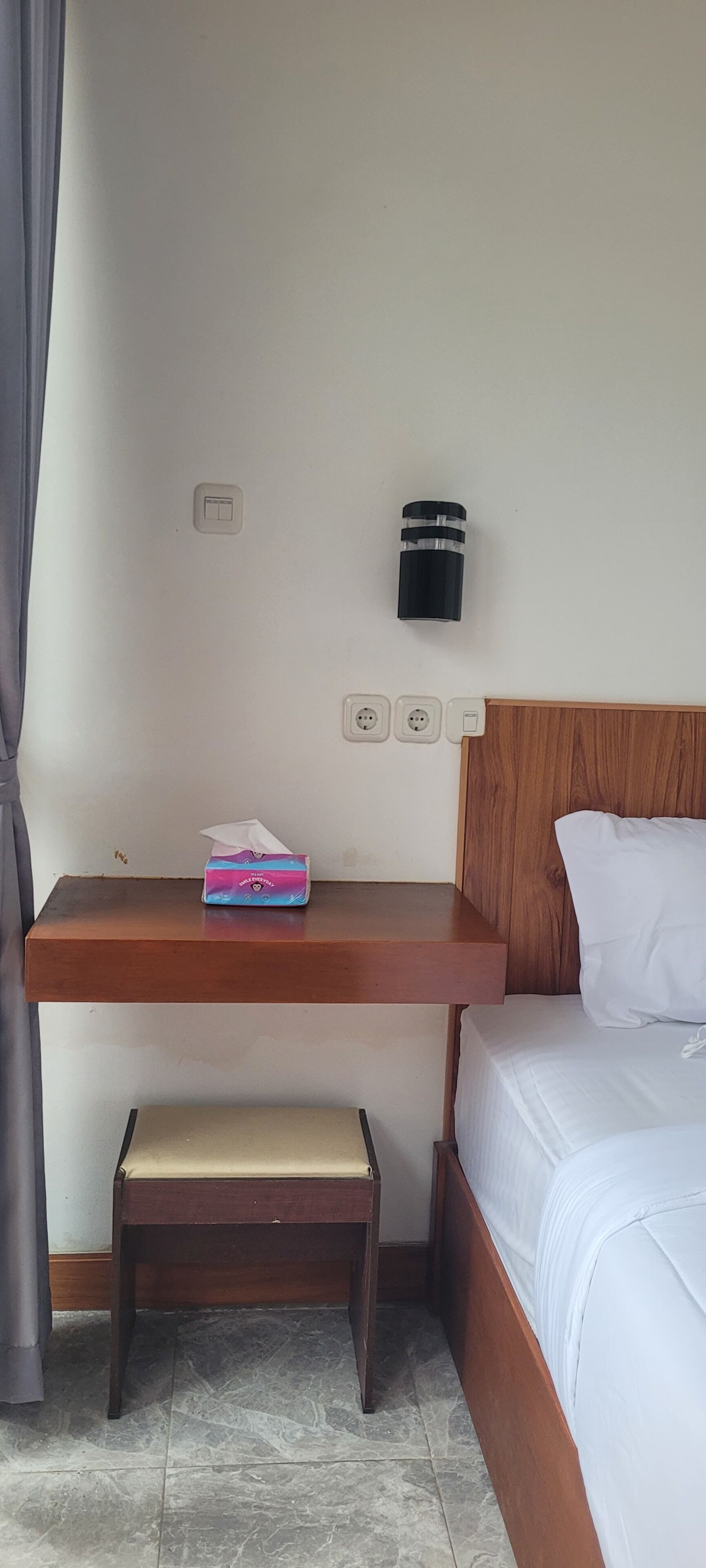 A hotel room with a wooden bed, a bedside table with a tissue box, a black thermos, and a small wooden bench.