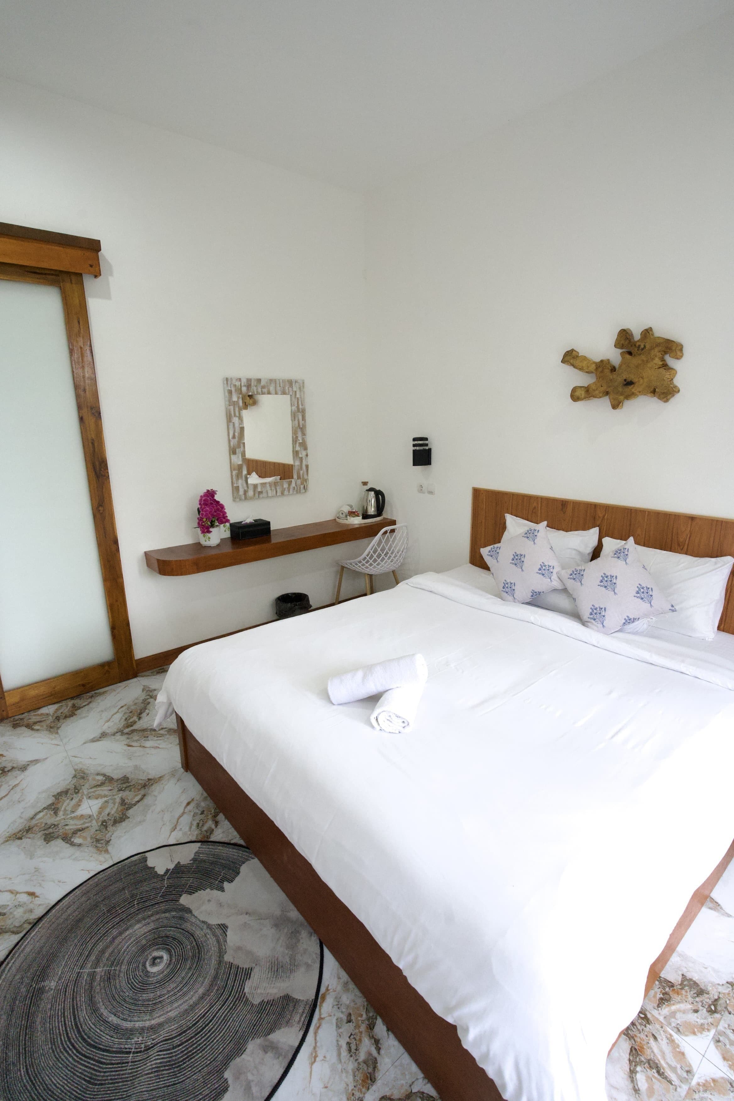 A neatly made bed with white linens and decorative pillows, a wooden headboard, a small shelf with a vase and a mirror, and a marble-patterned floor.