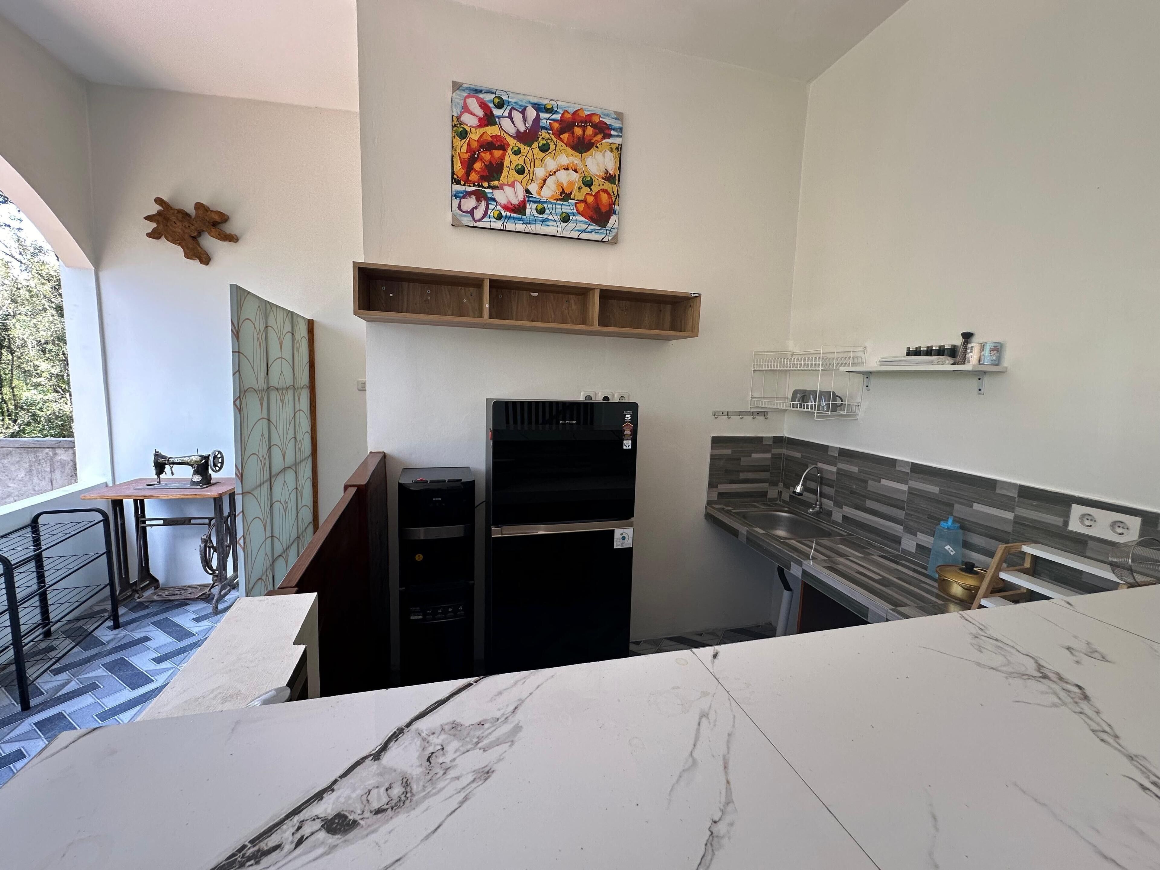 A compact kitchen with a marble countertop, a refrigerator, a sink, and a wooden shelf above the counter.