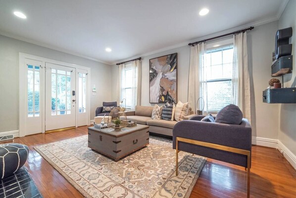 A spacious living room with a sofa, a coffee table, and a rug. There are large windows with curtains, a door, and a wall-mounted shelf with decorative items.