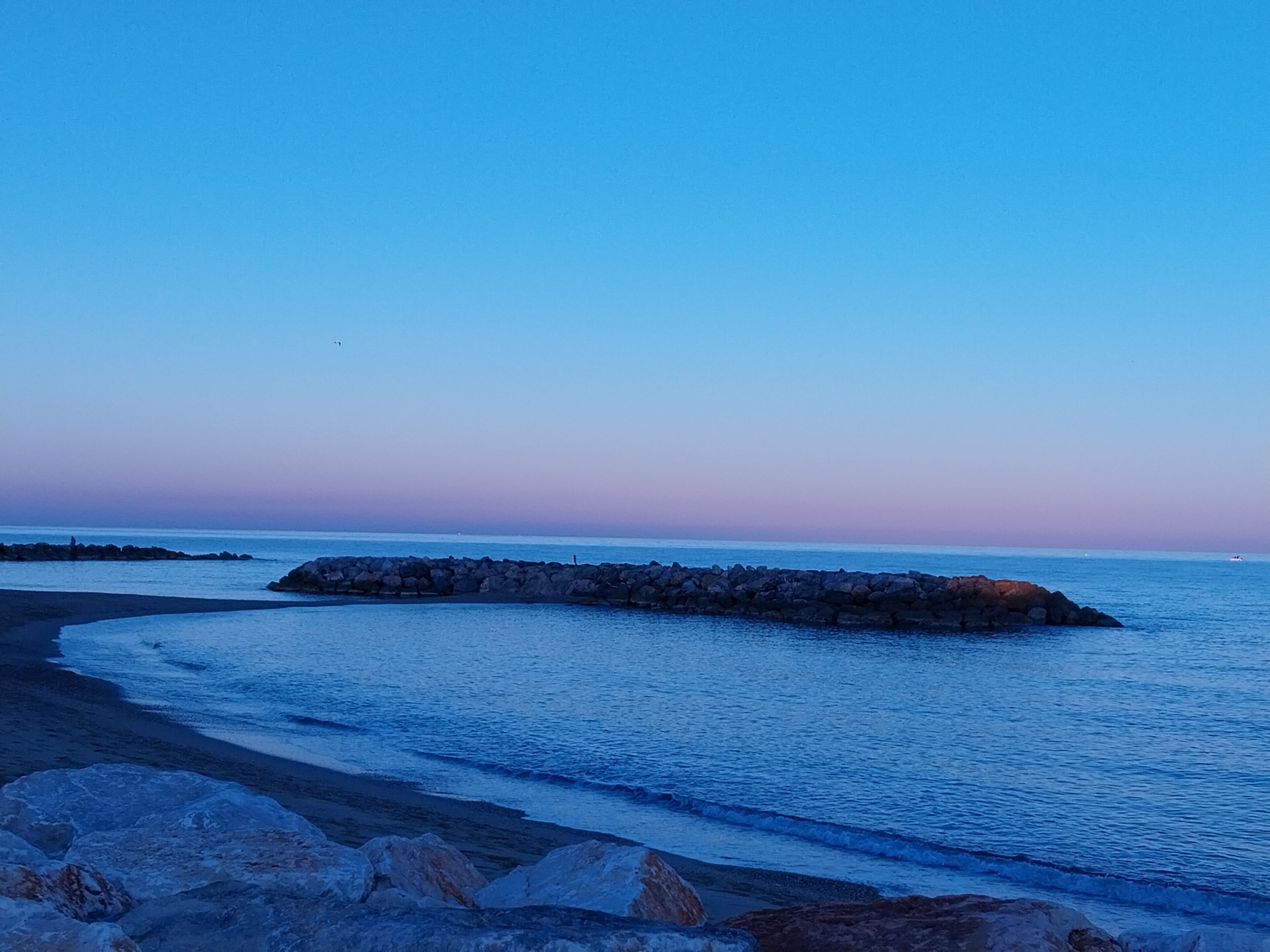 Un paysage côtier avec une digue rocheuse s’étendant dans des eaux calmes, un ciel dégagé et une plage de sable.