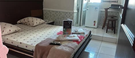 Habitación de hotel con cama, almohadas, un control remoto, un folleto y una pequeña zona de kitchenette.
