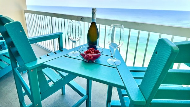 A table with a bottle, glasses, and a bowl of fruit on a balcony with a view of the ocean.