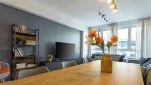 Un salon moderne avec une table à manger en bois, un téléviseur à écran plat, une bibliothèque et une grande fenêtre avec des rideaux.