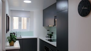 Une cuisine moderne avec des meubles de couleur sombre, un plan de travail blanc, un évier, une fenêtre avec des stores, une horloge et un mixeur.