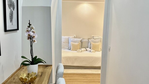 A modern, minimalist room with a wooden dining table, white upholstered chairs, a potted orchid, and a bed with white bedding in the background. - Charming apartment (Paris)