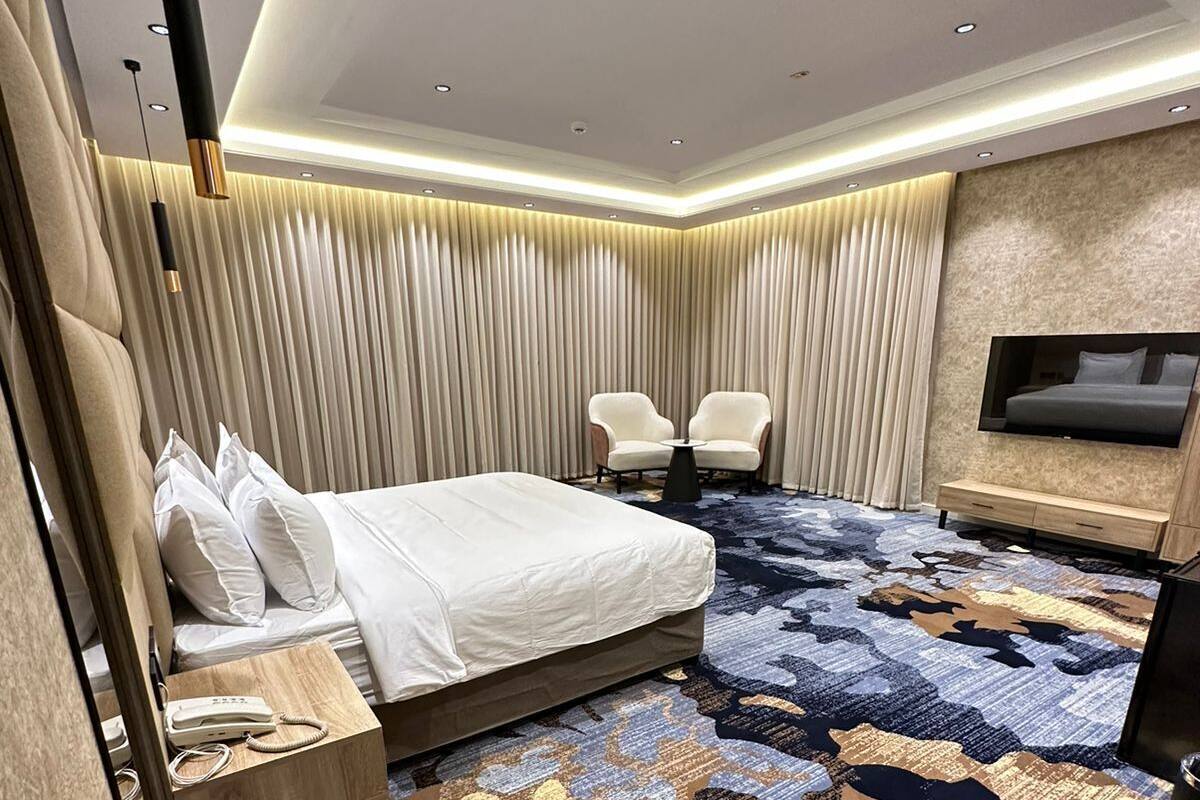 A modern hotel room with a large bed, a small seating area, a TV mounted on the wall, and a patterned carpet.