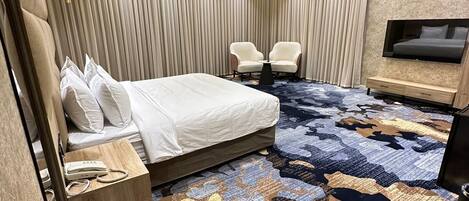 A modern hotel room with a large bed, a small seating area, a TV mounted on the wall, and a patterned carpet.