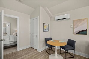 A hotel room with a wooden floor, a round table with two chairs, a door, an air conditioning unit, and a framed picture on the wall.