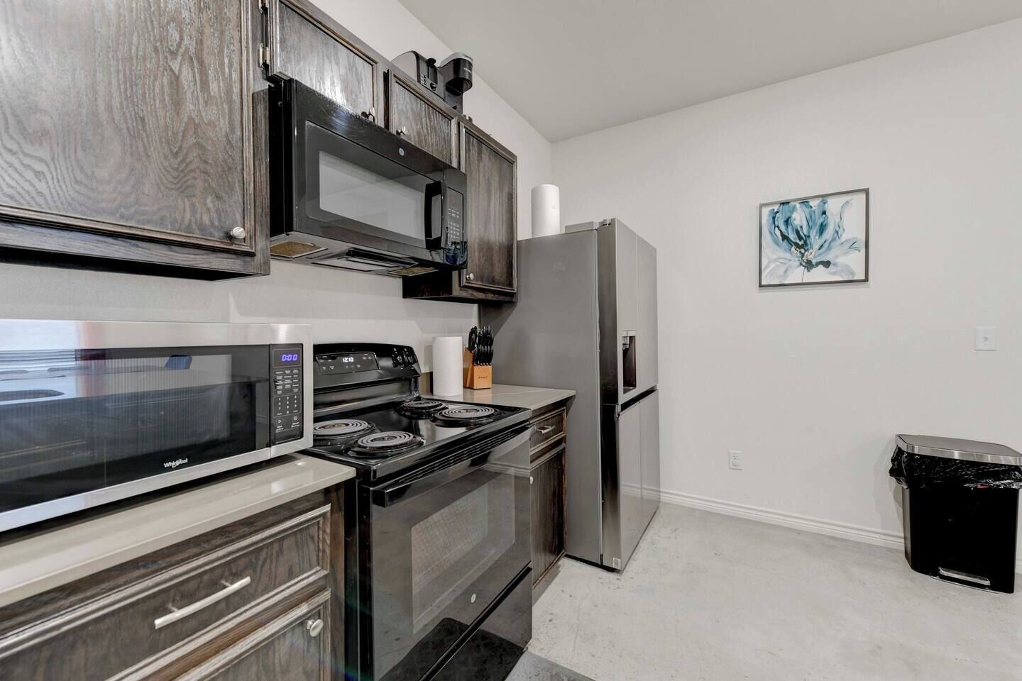 A kitchen with a stainless steel refrigerator, a microwave, an oven, and a stove. There is a trash bin and a painting on the wall.