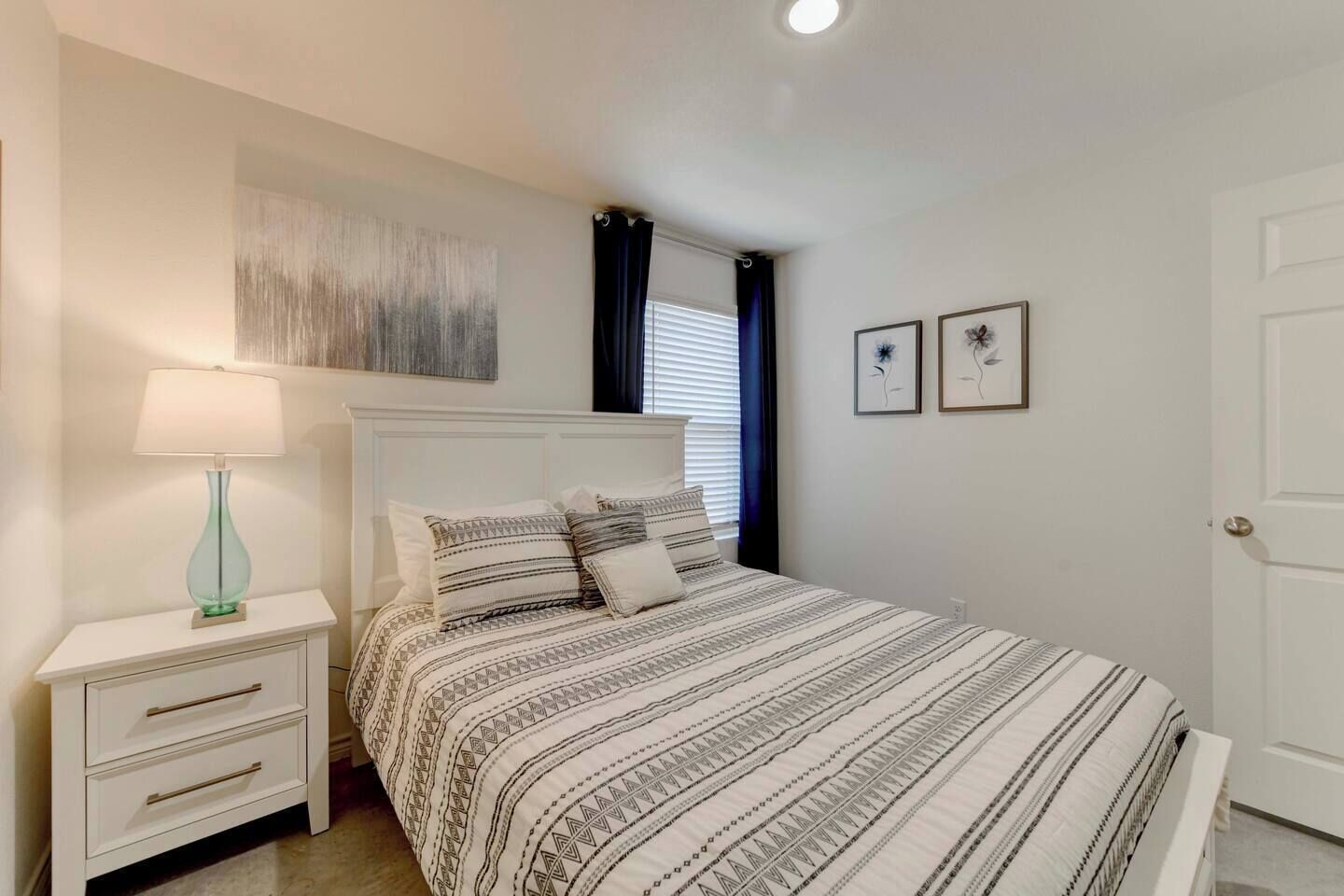 Un dormitorio con cama, mesitas de noche, lámpara, ventana con cortinas y dos cuadros en la pared.