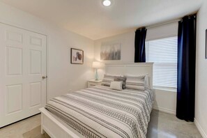 Un dormitorio con cama, mesitas de noche, una puerta, una ventana con cortinas y una obra de arte colgada en la pared.