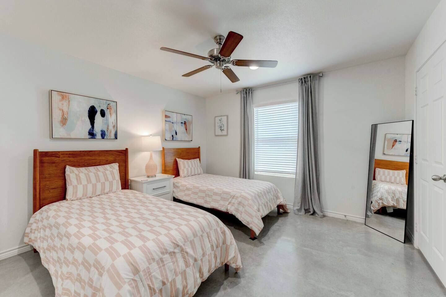 A bedroom with two beds, a ceiling fan, a window with blinds, and artwork on the walls.