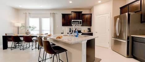 A modern kitchen with a central island, dark wood cabinets, stainless steel appliances, and a dining area with chairs and a table.