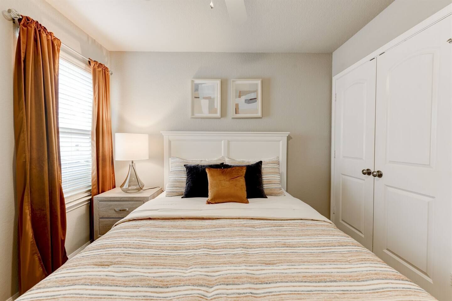 A bedroom with a bed, nightstand, lamp, two framed pictures on the wall, and a wardrobe.